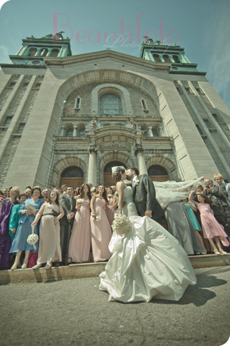 Bride and groom exchanging a kiss in front of their church with all their guests in the back